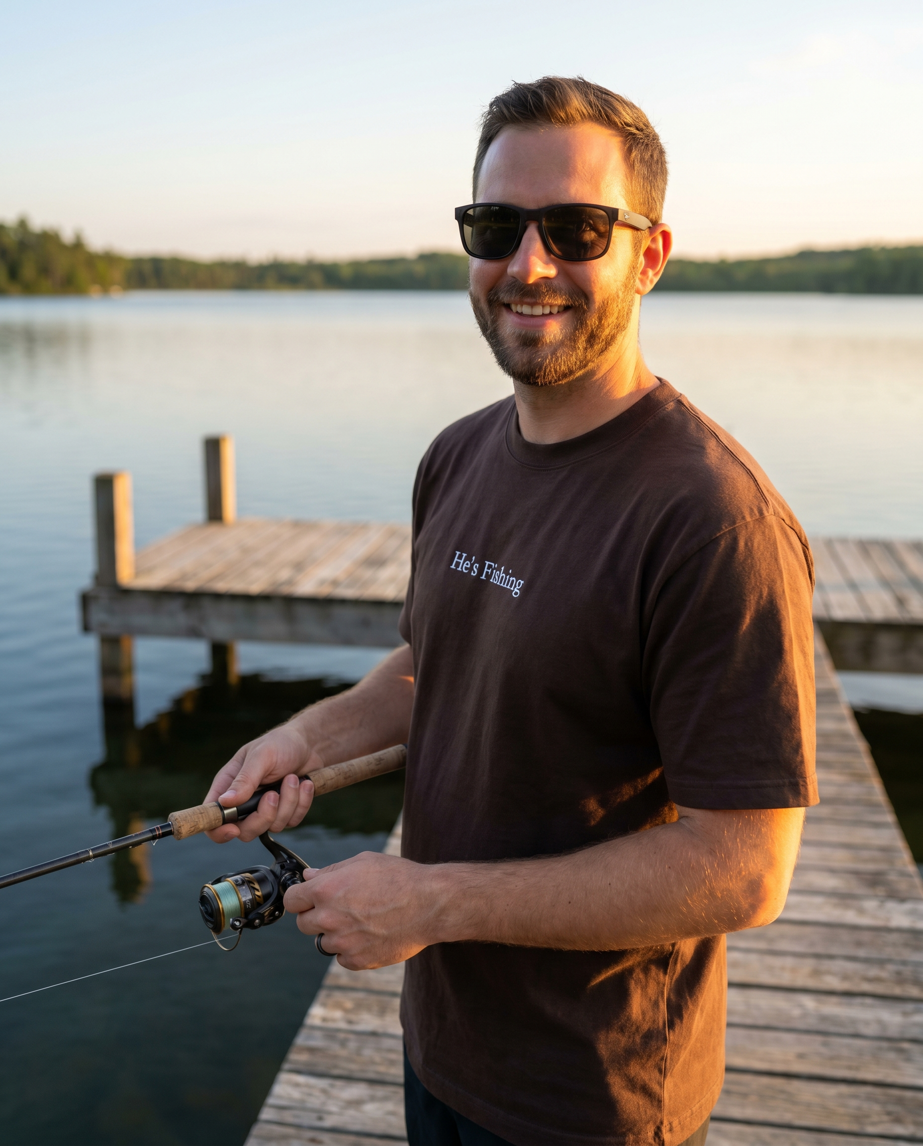 Man wearing He's Fishing espresso t-shirt holding a fishing rod on a dock at sunset