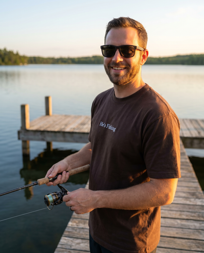 Man wearing He's Fishing espresso t-shirt holding a fishing rod on a dock at sunset