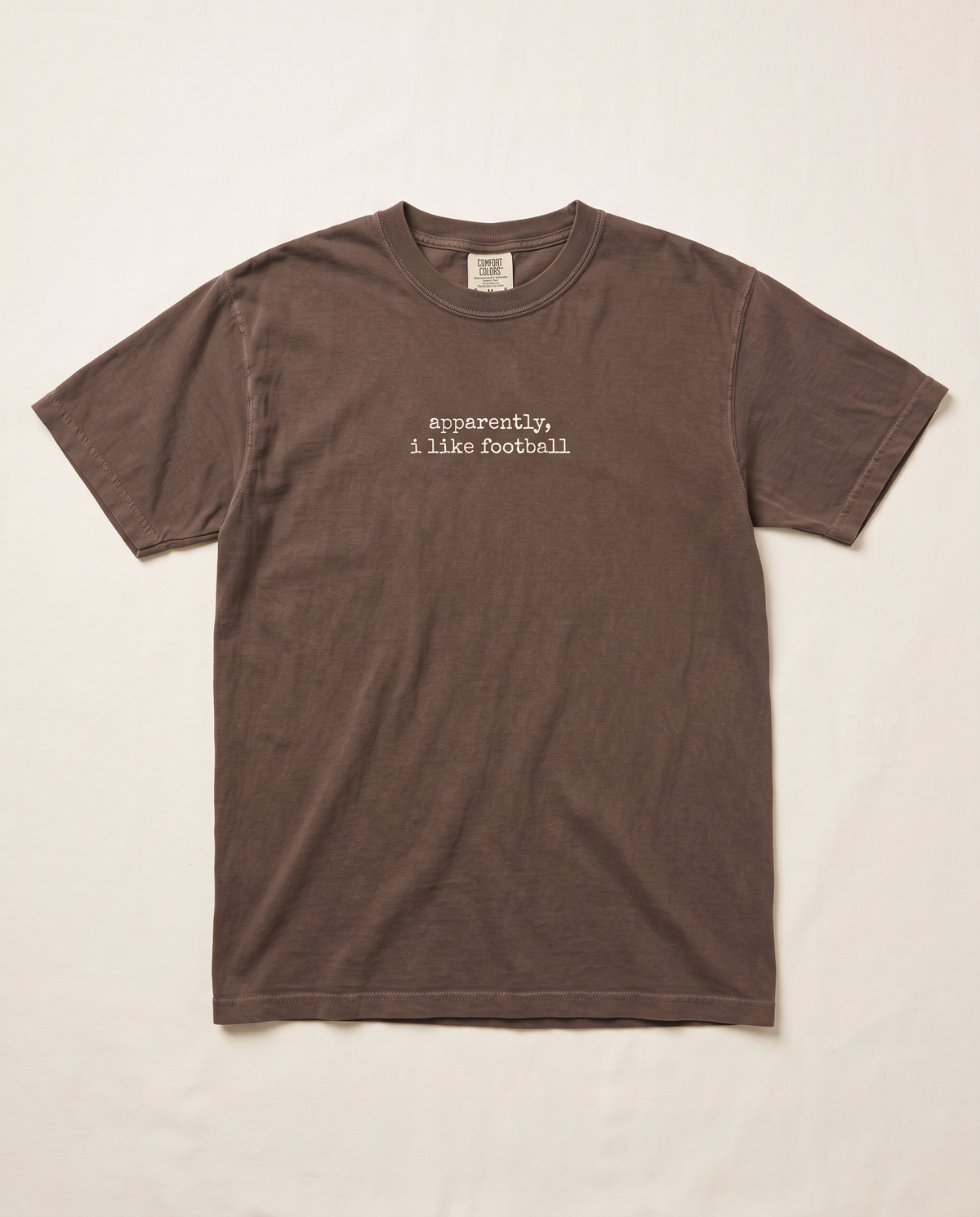 Apparently I Like Football tee — small cream chest print on espresso Comfort Colors tee, flat lay